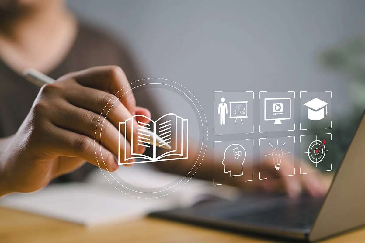 Mano che scrive accanto a icone digitali di apprendimento e materiali didattici su uno schermo laptop