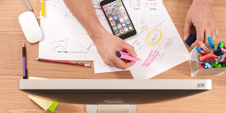 Persona evidenzia appunti e progetta contenuti digitali su una scrivania con computer, smartphone e pennarelli colorati