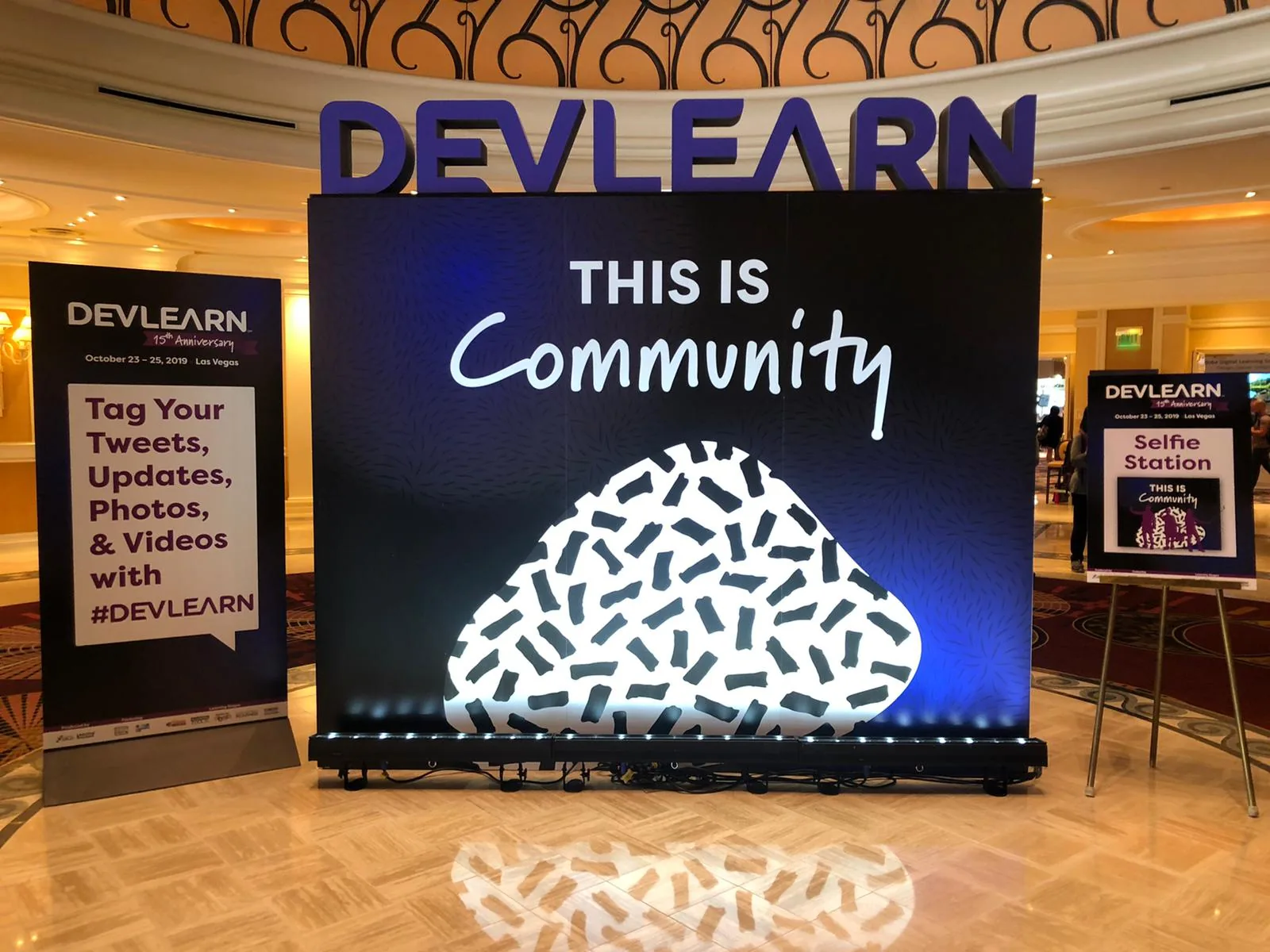 Allestimento DevLearn 2019 con pannello This is Community e indicazioni per social e selfie station