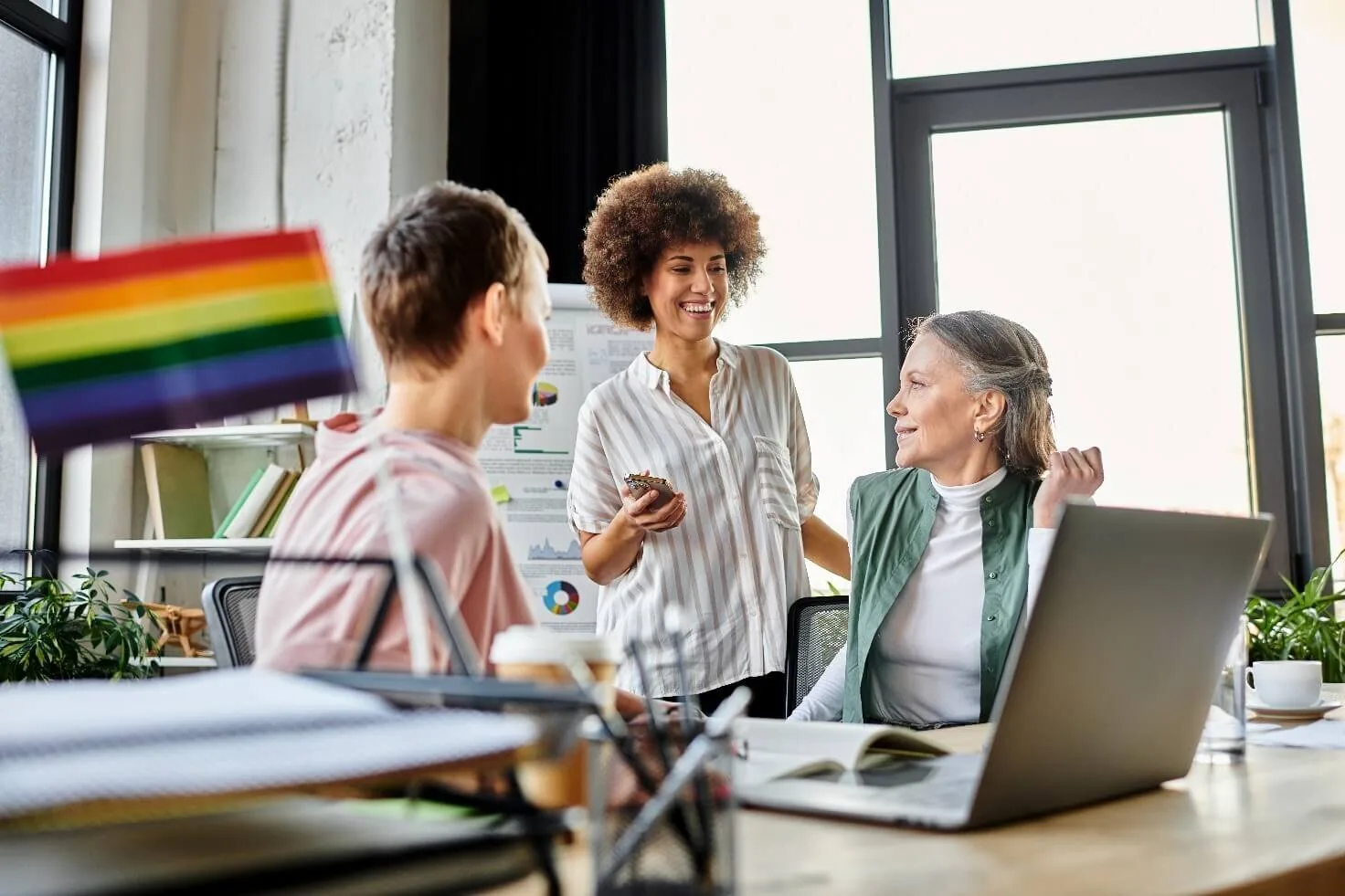 Tre persone di diverse età interagiscono in ufficio con una bandiera arcobaleno in primo piano, simbolo di inclusione