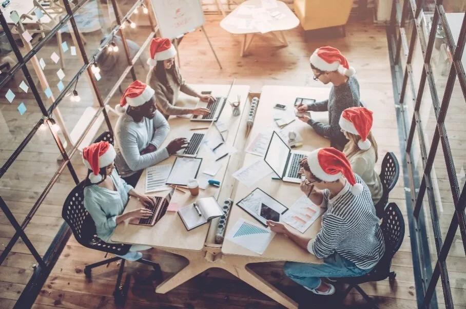 Gruppo di colleghi in ufficio con cappelli di Natale che lavorano insieme a un tavolo condiviso durante il periodo festivo