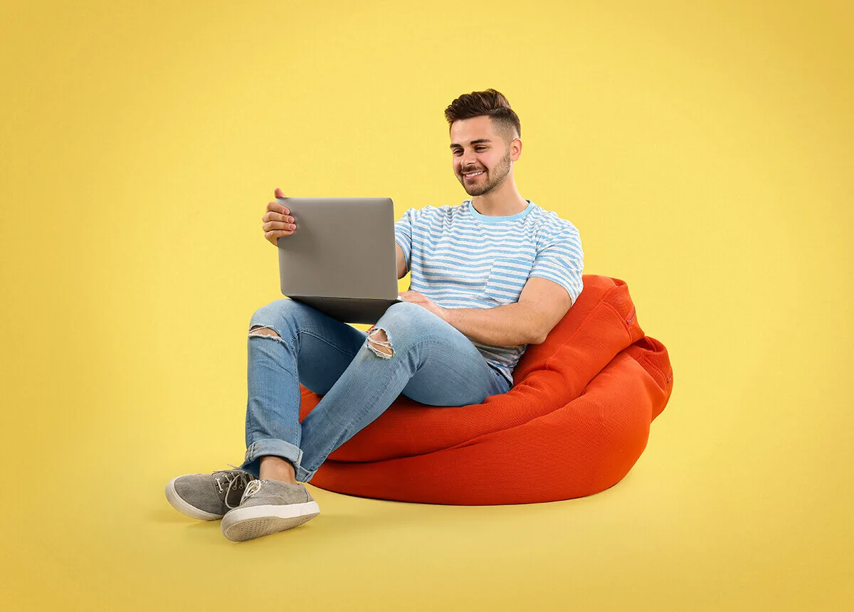Giovane uomo lavora da remoto seduto su pouf arancione con laptop su sfondo giallo, simbolo di smart working moderno
