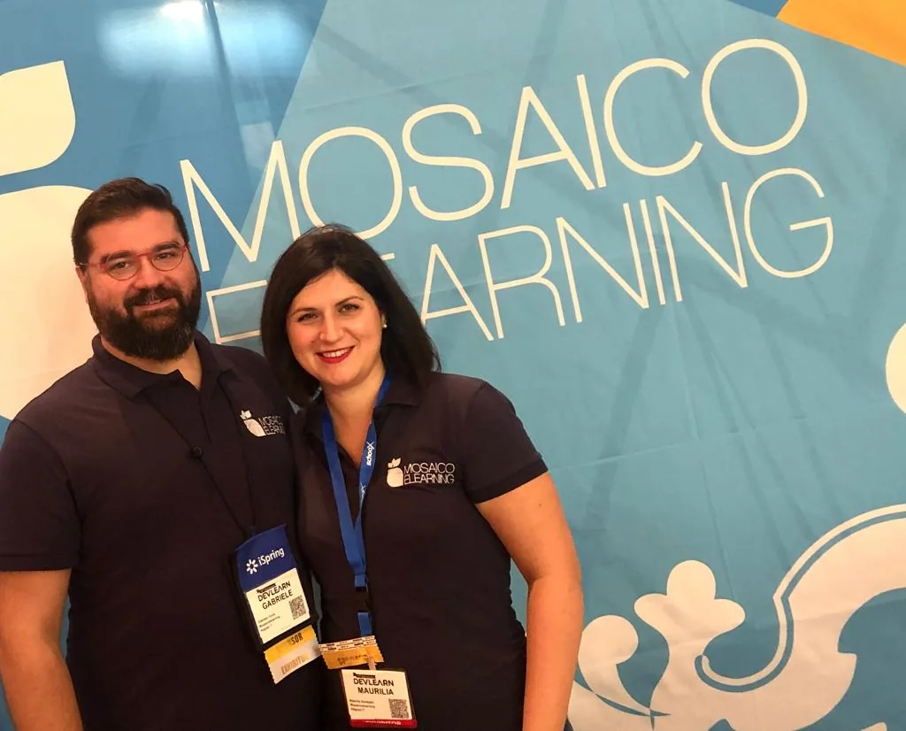 Due persone con polo Mosaico e badge DevLearn davanti a un banner Mosaico eLearning durante un evento