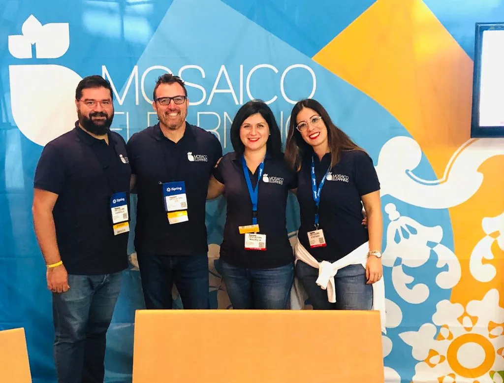 Quattro membri del team Mosaico eLearning sorridono davanti allo stand colorato durante l’evento DevLearn